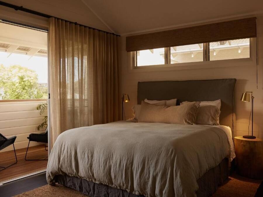 A bedroom with a large bed, a window with curtains, a bedside table with a lamp, and a chair by the window.
