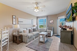 A living room with a beige sofa, a wooden coffee table, and a ceiling fan. There is a window with a view of greenery outside. - Gorgeous Ocean View - Grande Caribbean Orange Beach 2 BR Condo (Orange Beach)