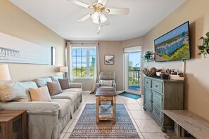 A living room with a sofa, a wooden coffee table, a ceiling fan, a TV mounted on the wall, and a view of the outdoors through a window. - Gorgeous Ocean View - Grande Caribbean Orange Beach 2 BR Condo (Orange Beach)