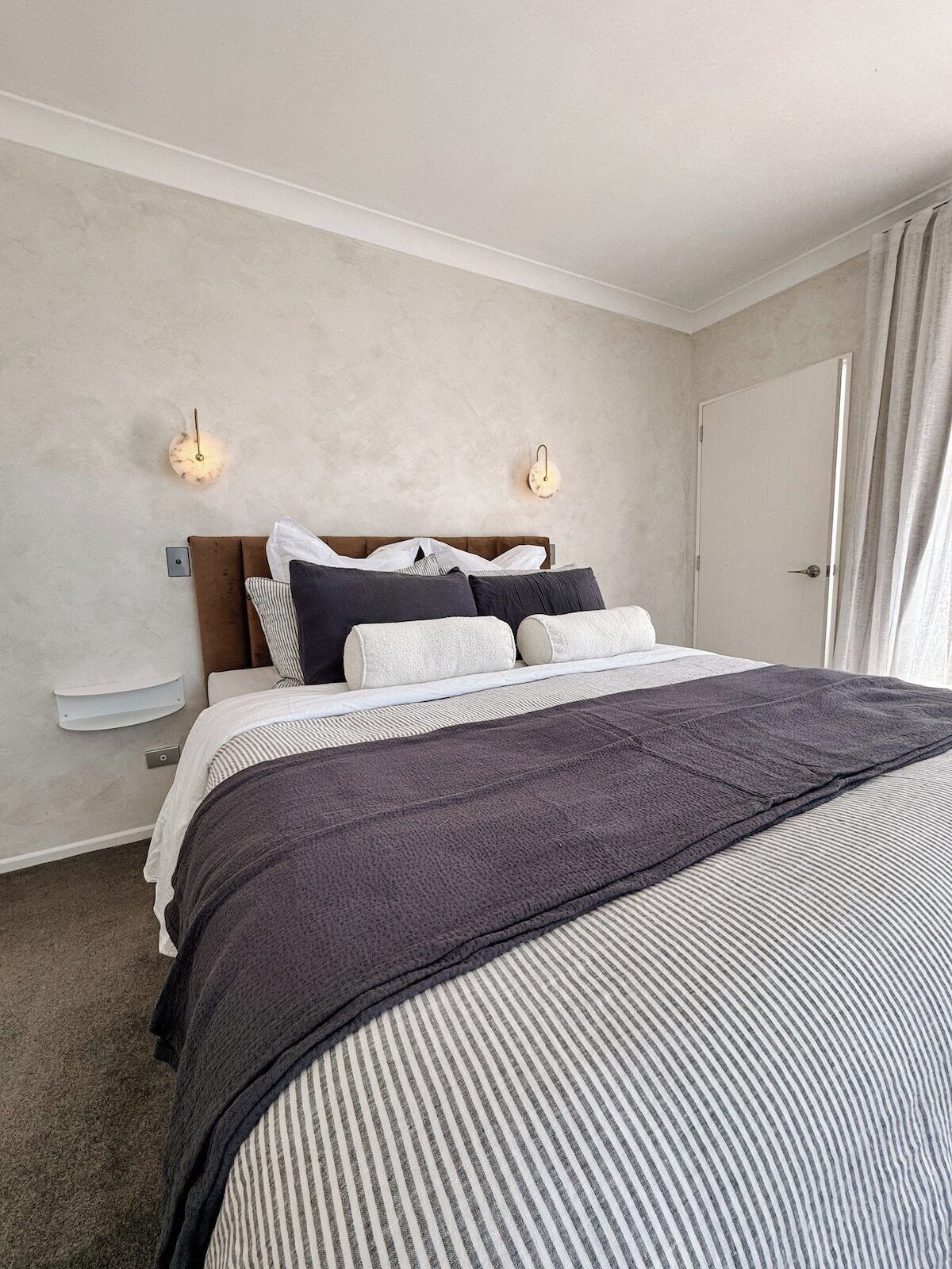 Ein Schlafzimmer mit Bett, Kopfteil, Nachttischen, Wandlampen und einer Tür.