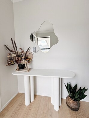 A white console table with a mirror above it, a potted plant to the right, and a decorative arrangement on the left.
