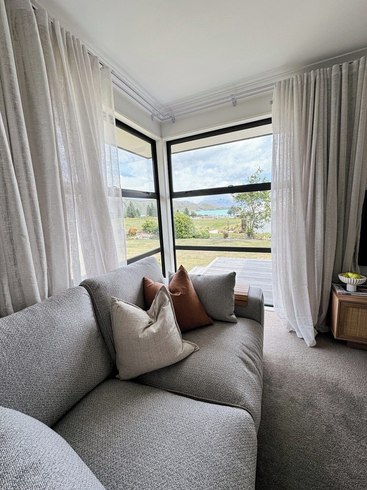 A modern living room with a grey sofa, white curtains, and a view of a landscape through large windows.