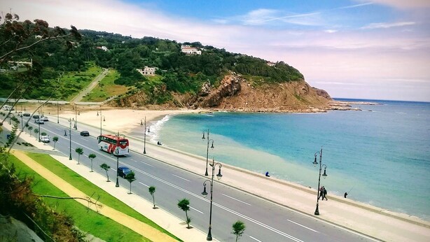 Una carretera costera con una playa a un lado y una zona montañosa con edificios al otro.