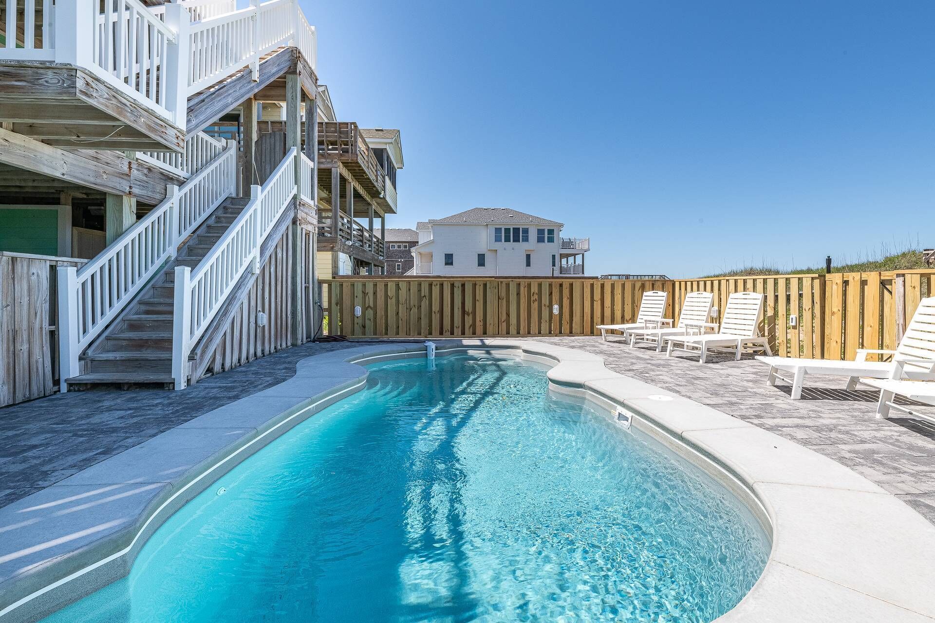 Ein Schwimmbad mit Holzterrasse und Liegestühlen, umgeben von einem Holzzaun und einem Gebäude mit mehreren Balkonen.