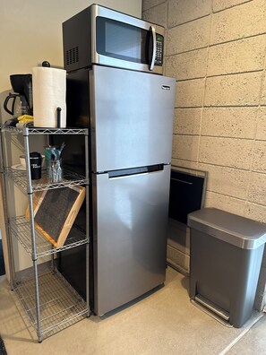 Ein Edelstahl-Kühlschrank mit einer Mikrowelle obenauf, einer kleinen Mülltonne und einem Metallregal mit Küchenutensilien.