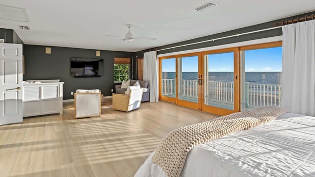 A modern bedroom with a large bed, a seating area with a sofa and armchair, a TV, and a balcony with a view of the ocean. - Walks on the Shores • Private Beach Front Access (Indian Shores)
