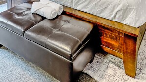 Un dormitorio con cabecera de madera, una cama grande con muchos almohadones, una mesita de noche con lámpara, y un banco con toallas.