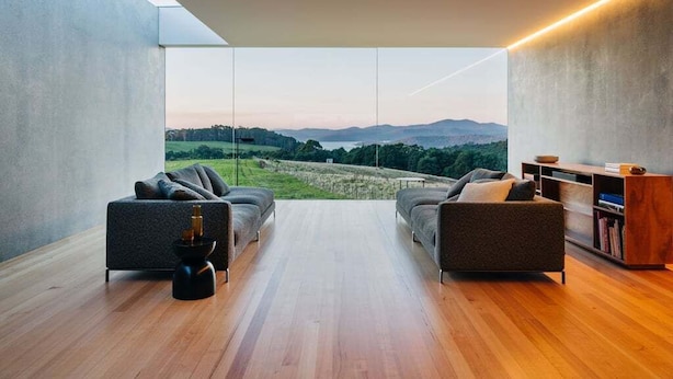 A modern living room with a large window overlooking a scenic landscape, featuring two sofas and a wooden floor. - Triptych - The Blunt House (Tasman)
