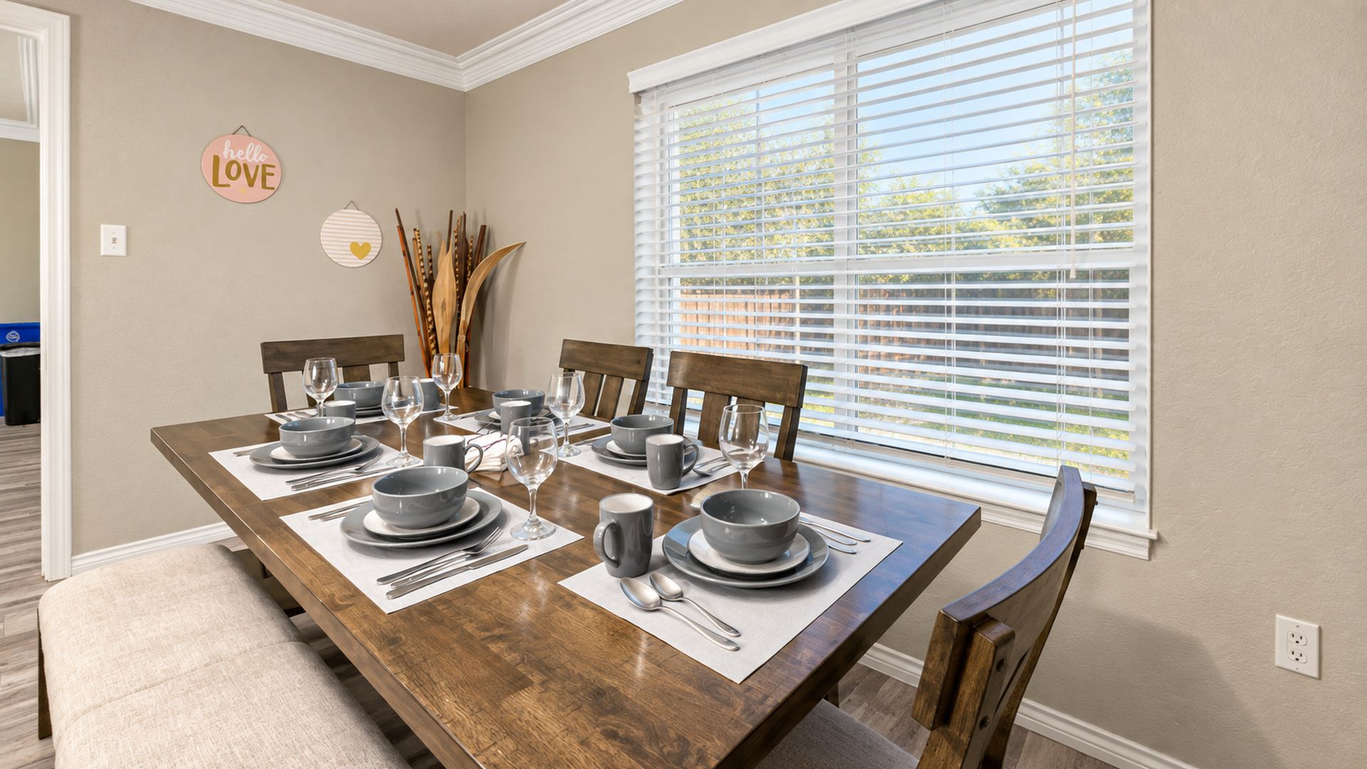 A dining room with a wooden table set for four, surrounded by chairs, with a window with blinds and a wall decoration.