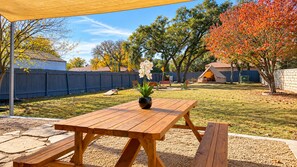 An outdoor patio with a wooden table and benches, surrounded by a backyard with a wooden fence, trees, and a tent. - Glampstead Retreat | BMT|SixFlags|SeaWorld|Cantera (San Antonio)