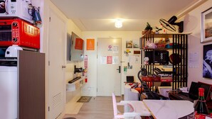 Et kompakt køkken med moderne hårde hvidevarer, et spiseområde med bord og stole, og et lille opholdsområde med en reol.