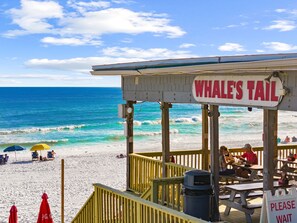 Een restaurant aan het strand met een bord waarop 'Whale's Tail' staat, zitplaatsen buiten en uitzicht op de oceaan.