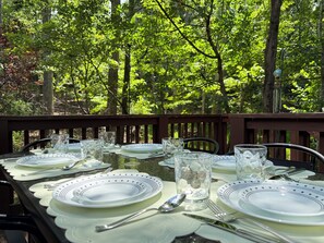 Une table dressée à l’extérieur, avec des assiettes, des verres et des couverts, entourée d’une zone boisée.