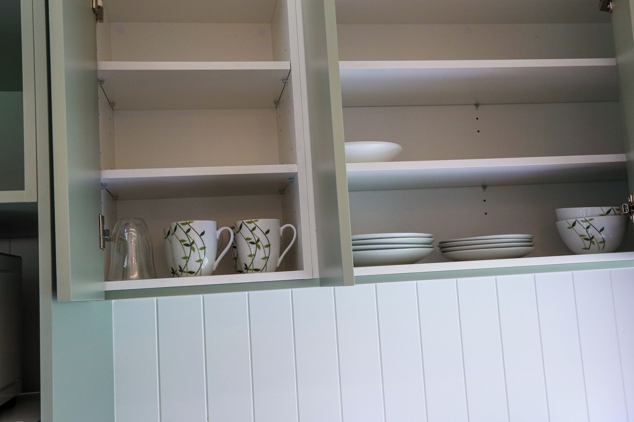 A kitchen with white cabinets and shelves, containing white dishes and cups with green leaf patterns.