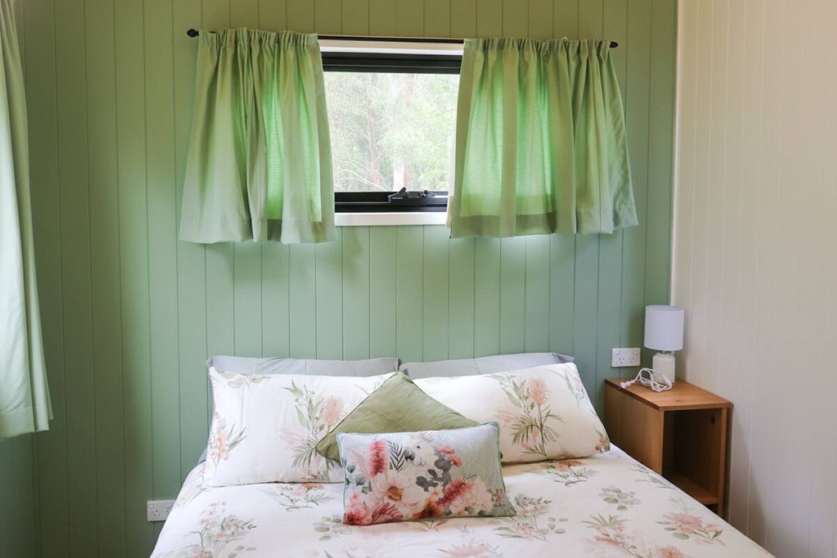 Un dormitorio con cama, ropa de cama estampada con motivos florales, una mesita de noche con lámpara y cortinas verdes.