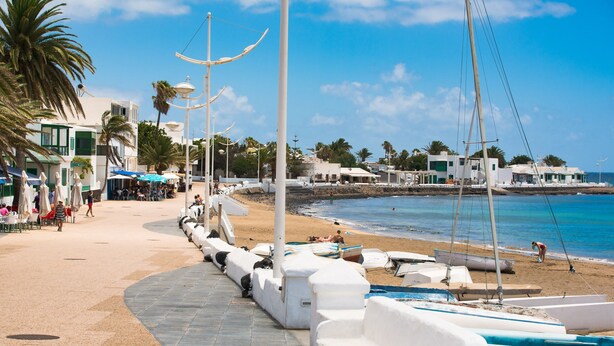 Un paseo marítimo con bancos blancos, una playa arenosa y barcos.