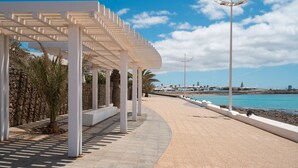 Un paseo marítimo con un pérgola blanca, palmeras y vistas al mar y a los barcos.