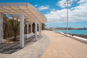Ein Küstenweg mit einer weißen Pergola, Palmen und Blick auf das Meer und Boote.