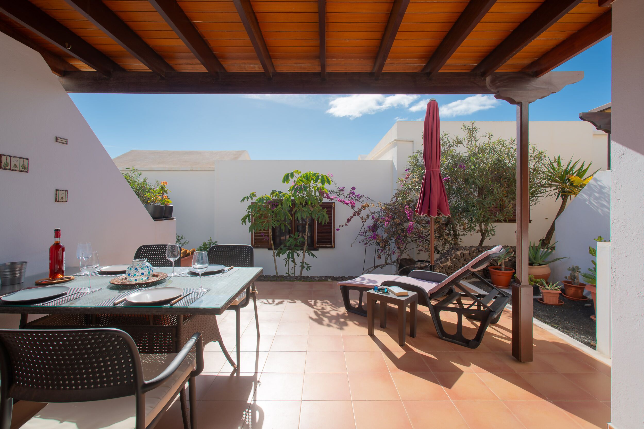 Un patio al aire libre con un pergolado de madera, una mesa de comedor preparada con platos, vasos y cubiertos, rodeada de sillas y macetas con plantas.