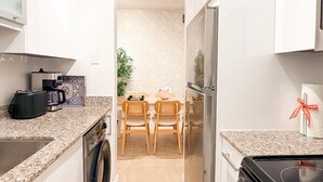 Eine moderne Küche mit Waschmaschine, Kühlschrank und Herd. Im Hintergrund ist ein Essbereich mit zwei Stühlen und einem Tisch zu sehen.
