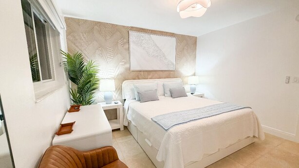 Ein Schlafzimmer mit einem Bett, Nachttischen, einem Sessel, einer Topfpflanze und einem Wandbild.