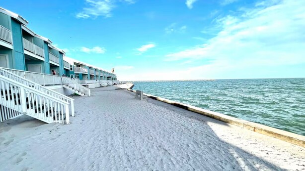 A beachfront hotel with a sandy walkway leading to the sea, a pier in the distance, and a clear blue sky. - Beachfront Water View in Perfect Paradise at Bayfront Kontiki! (Rockport)