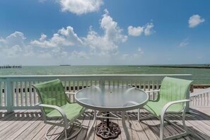 A deck with a round table and two chairs, overlooking a calm sea and a pier in the distance. - Beachfront Water View in Perfect Paradise at Bayfront Kontiki! (Rockport)
