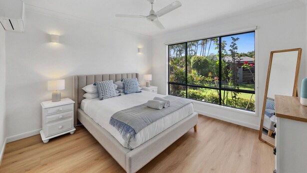 Um quarto com uma cama grande, um ventilador de teto, abajures, uma mesa de cabeceira, uma janela com vista para o verde e um espelho.