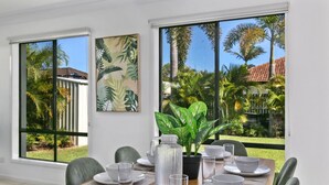 Uma área de refeições com uma mesa posta para quatro pessoas, grandes janelas com vista para palmeiras e uma casa, e um quadro na parede.