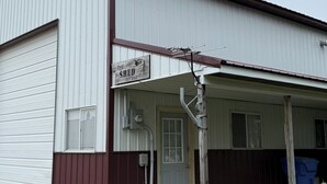 Un edificio de una planta con porche cubierto, un letrero que dice “The Shed” y una puerta con ventanal.