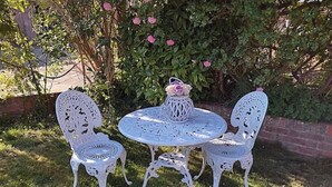 Een wit zitgedeelte buiten met een ronde tafel en twee stoelen, omringd door groen en een bakstenen muur.