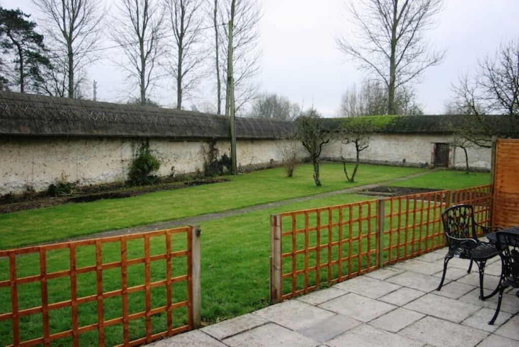 A courtyard with a thatched-roof building, a paved pathway, and a fenced area with chairs.