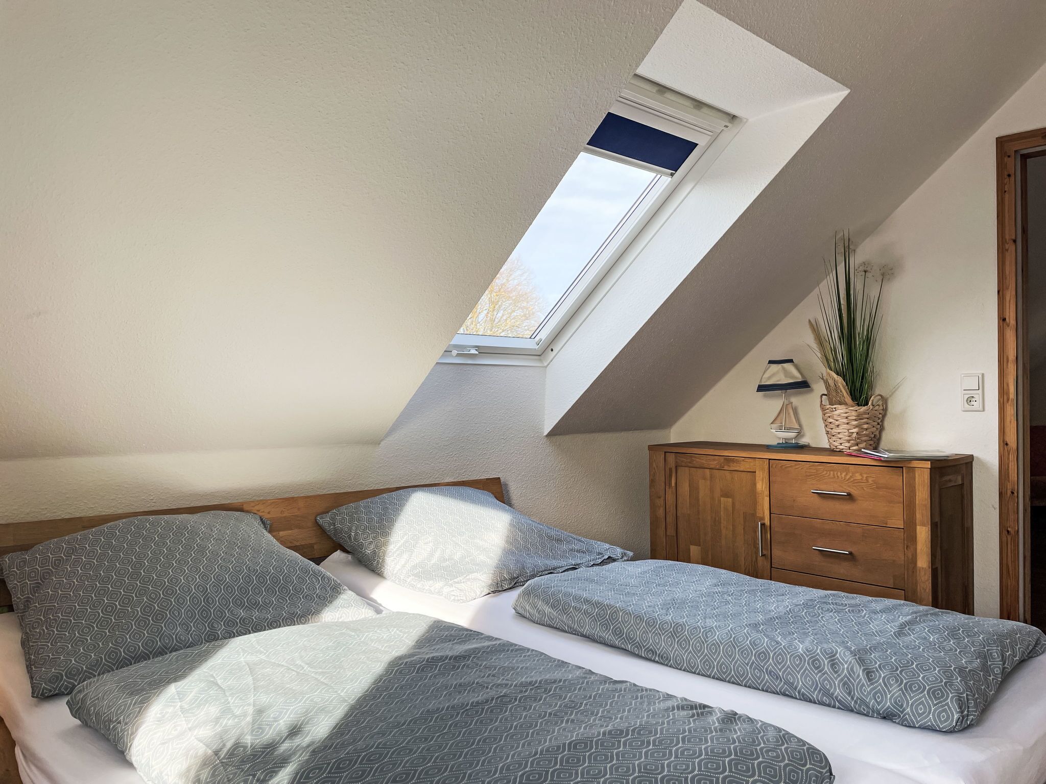 A bedroom with a sloped ceiling, a skylight, a bed with patterned bedding, a wooden dresser, and a potted plant.