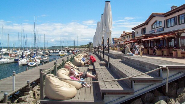 Een houten steiger met zitgedeeltes en parasols, met uitzicht op een jachthaven met boten.