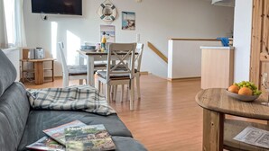 Moderni olohuone, jossa on harmaa sohva, ruokapöytä tuoleineen, seinälle asennettu televisio sekä keittiöalue, jossa on puukalusteet ja hedelmäkulho.