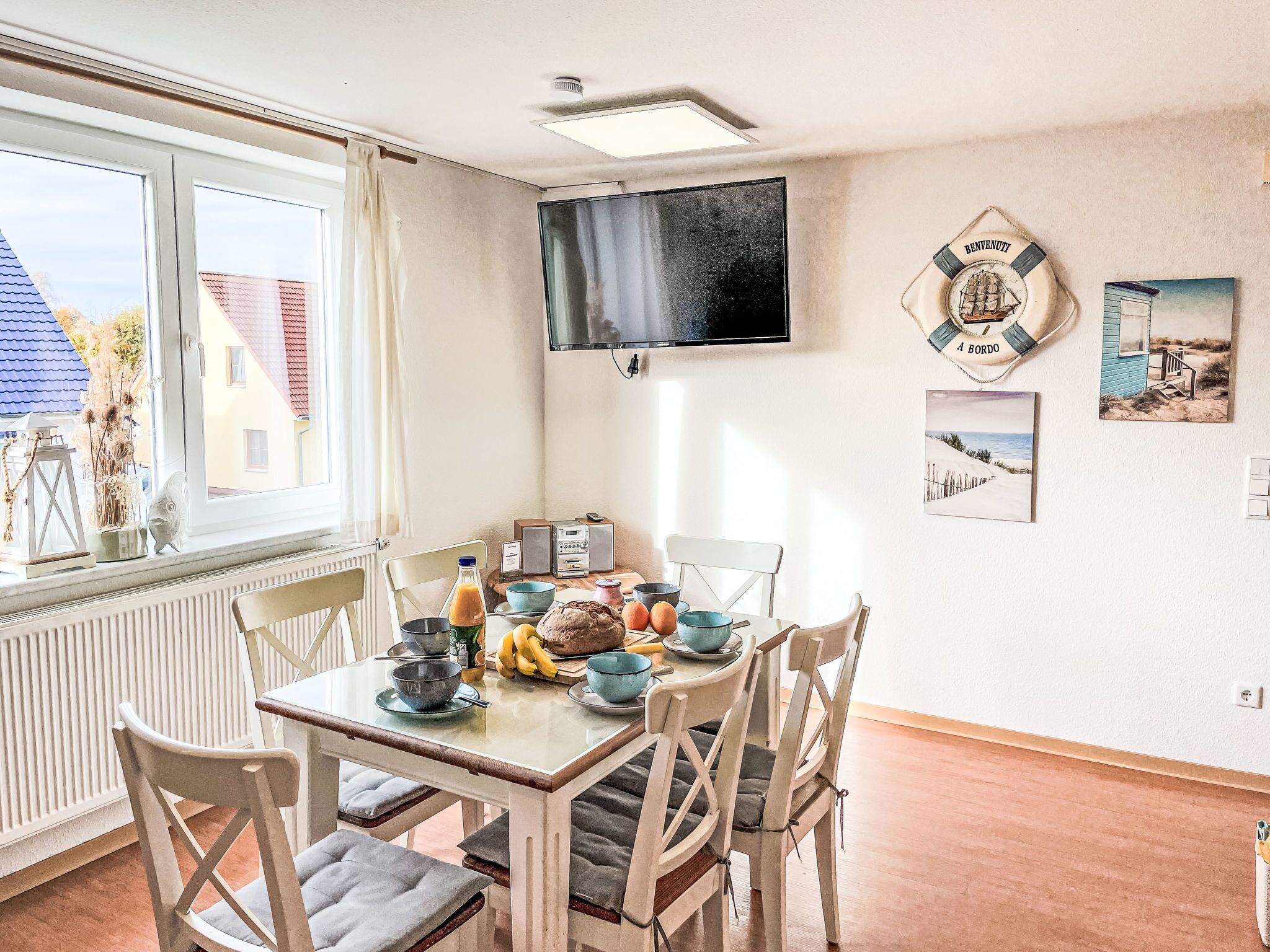 Un comedor con una mesa dispuesta para cuatro personas, un televisor montado en la pared y una ventana con vistas a casas.