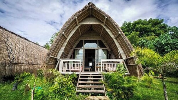 Ein traditionelles Gebäude mit einem Reetdach und einer zentralen Treppe, die zu einer Tür führt, umgeben von üppiger Vegetation.