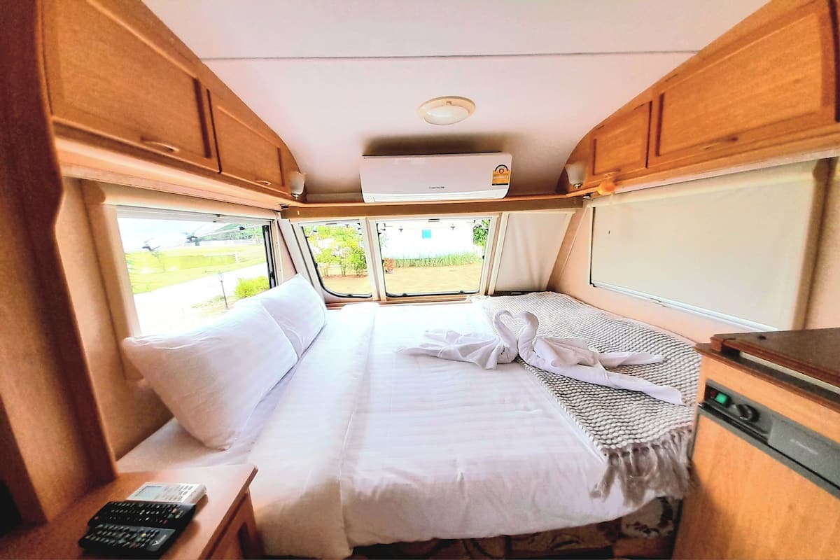 Interior compacto de una autocaravana con cama, una mesita, un mando a distancia y ventana con vistas al exterior.