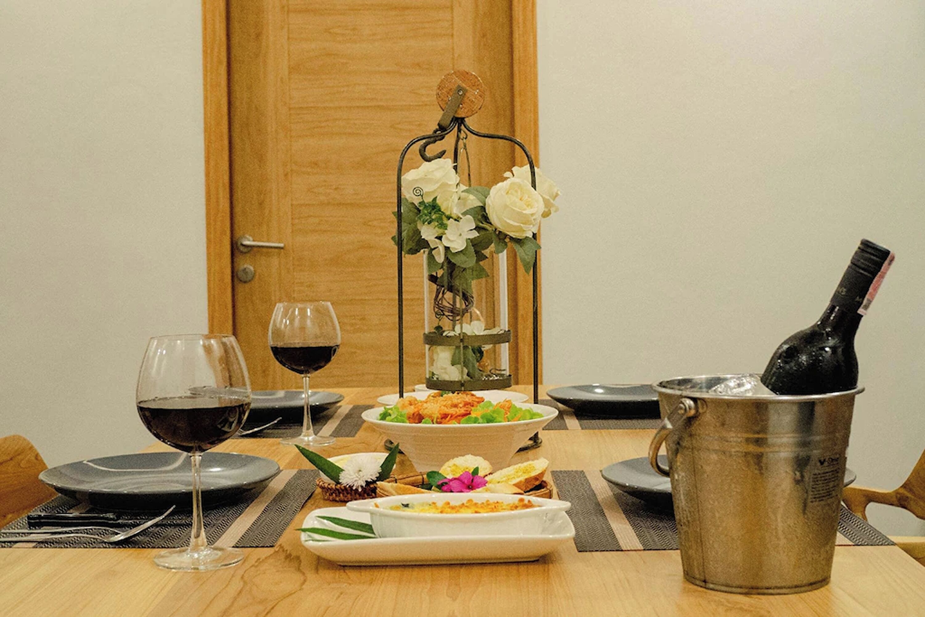 Une table à manger dressée avec des assiettes, des verres, une bouteille de vin, un seau en métal et un vase rempli de fleurs.