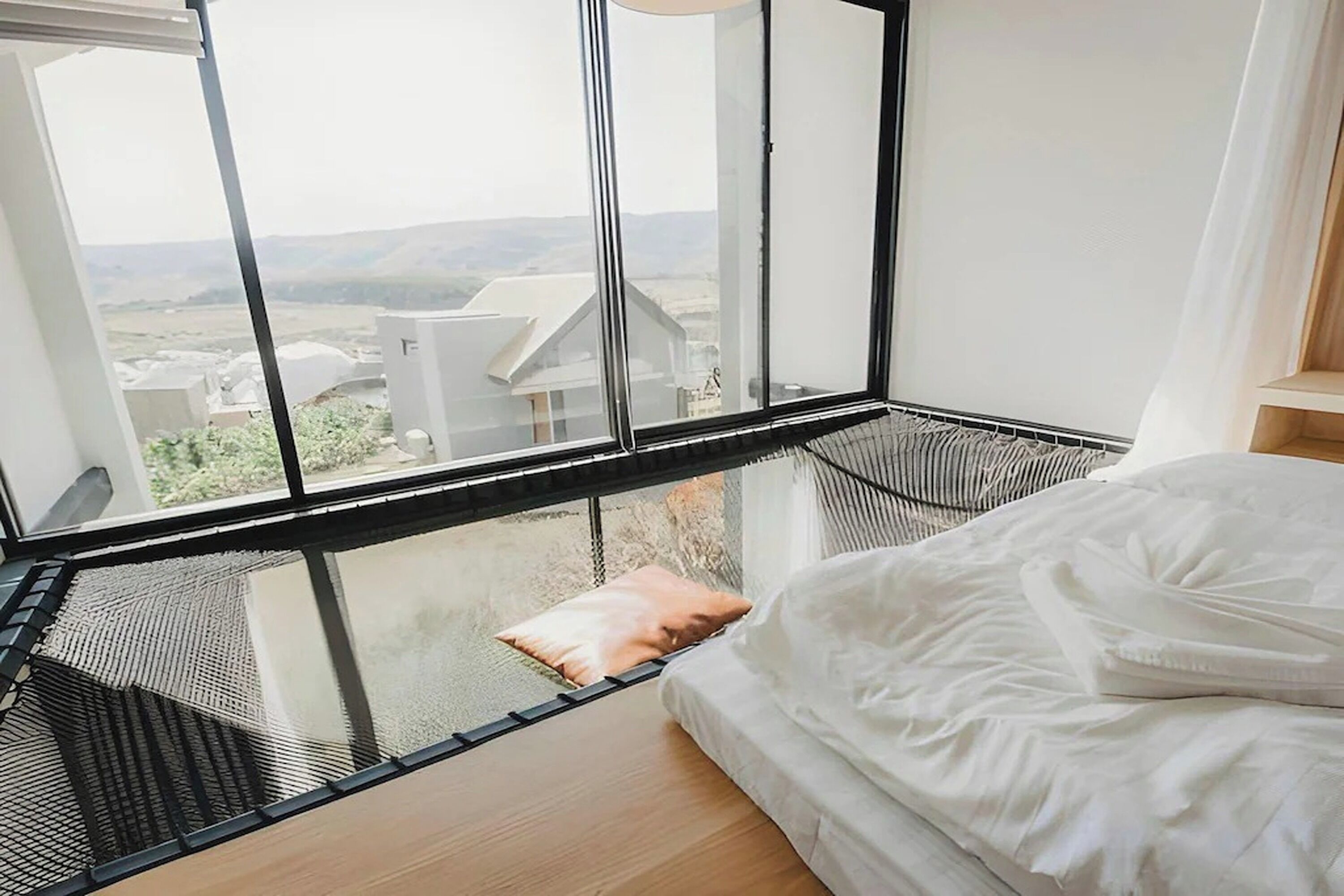 A bedroom with a large window, a bed, and a view of buildings and hills.