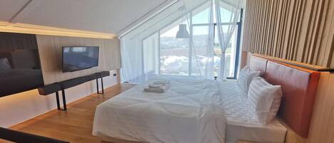 Un dormitorio moderno con una cama grande, un televisor en la pared y vistas al exterior a través de un amplio ventanal.