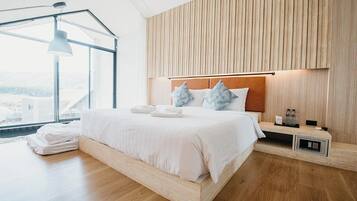 A modern bedroom with a wooden headboard, a large bed with white bedding, a bedside table with a lamp, and a view of the outdoors through large windows.