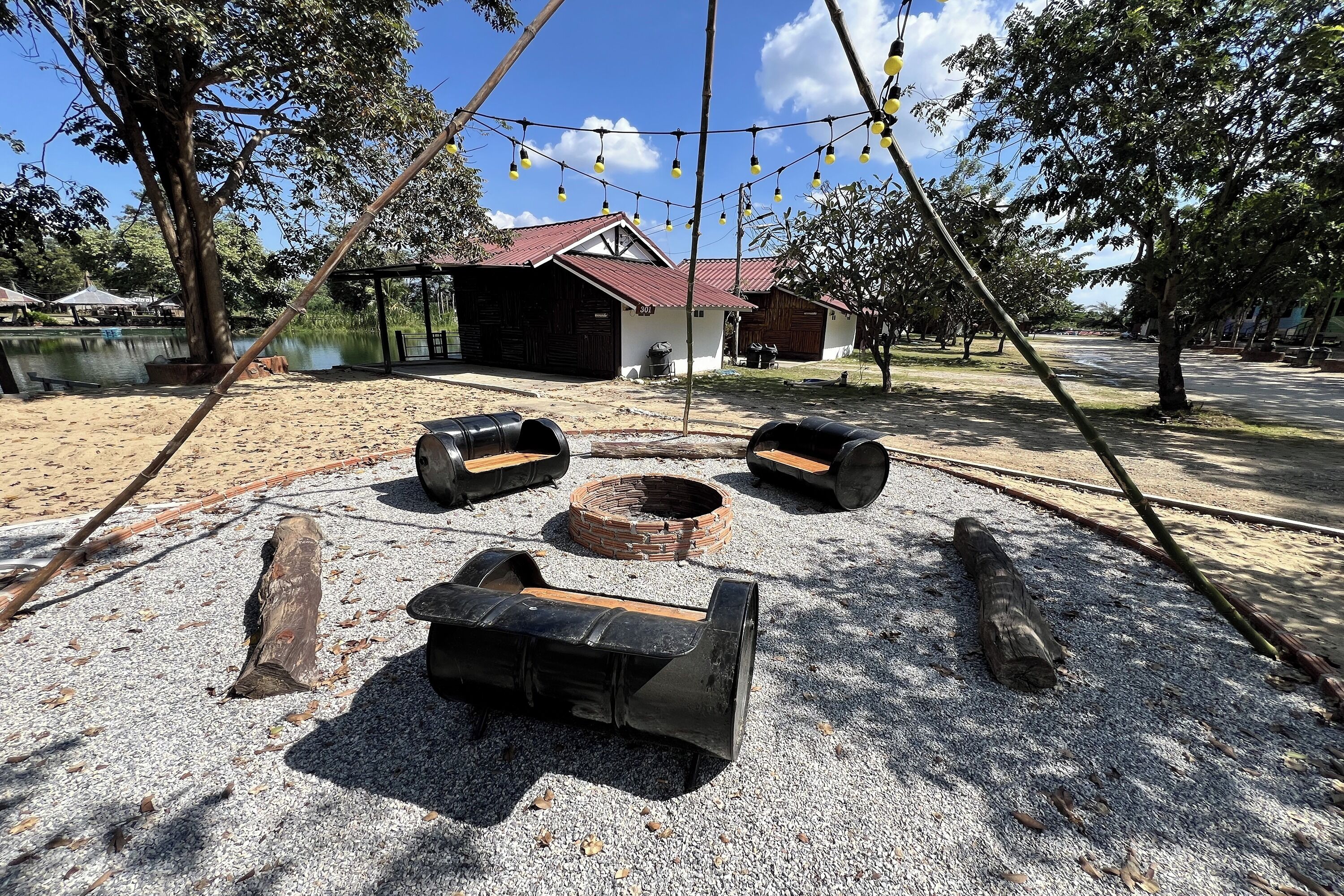 A rustic outdoor seating area with logs and benches, a small fire pit, and a building with a red roof in the background.