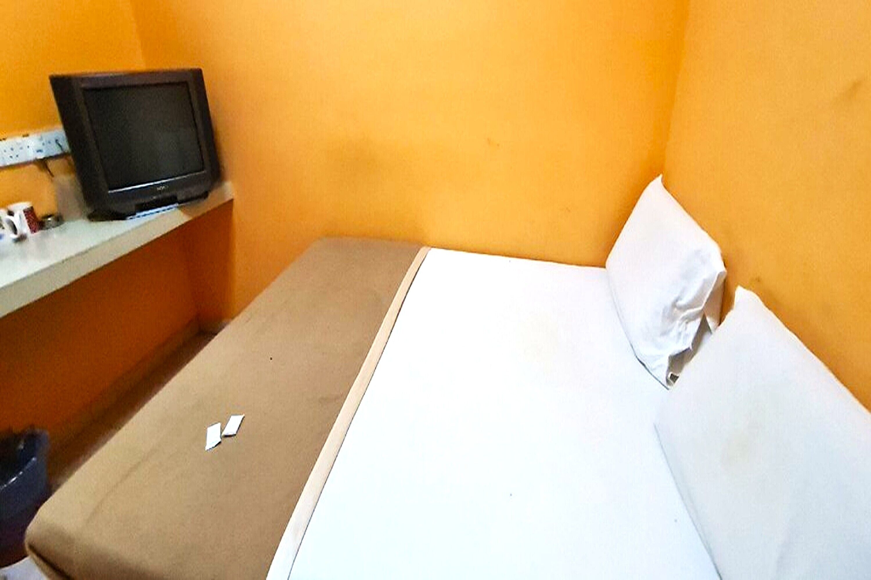 A small, single-bed room with a television on a shelf, a white pillow, and a beige bedspread.