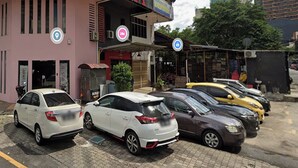 Une rangée de voitures garées devant un immeuble à la façade rose, sur lequel on peut voir une enseigne avec un « A » rouge.