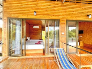 A wooden cabin with a balcony, a bed with white bedding, and a striped beach chair. - Mataburi River Kwai Resort (Sai Yok)