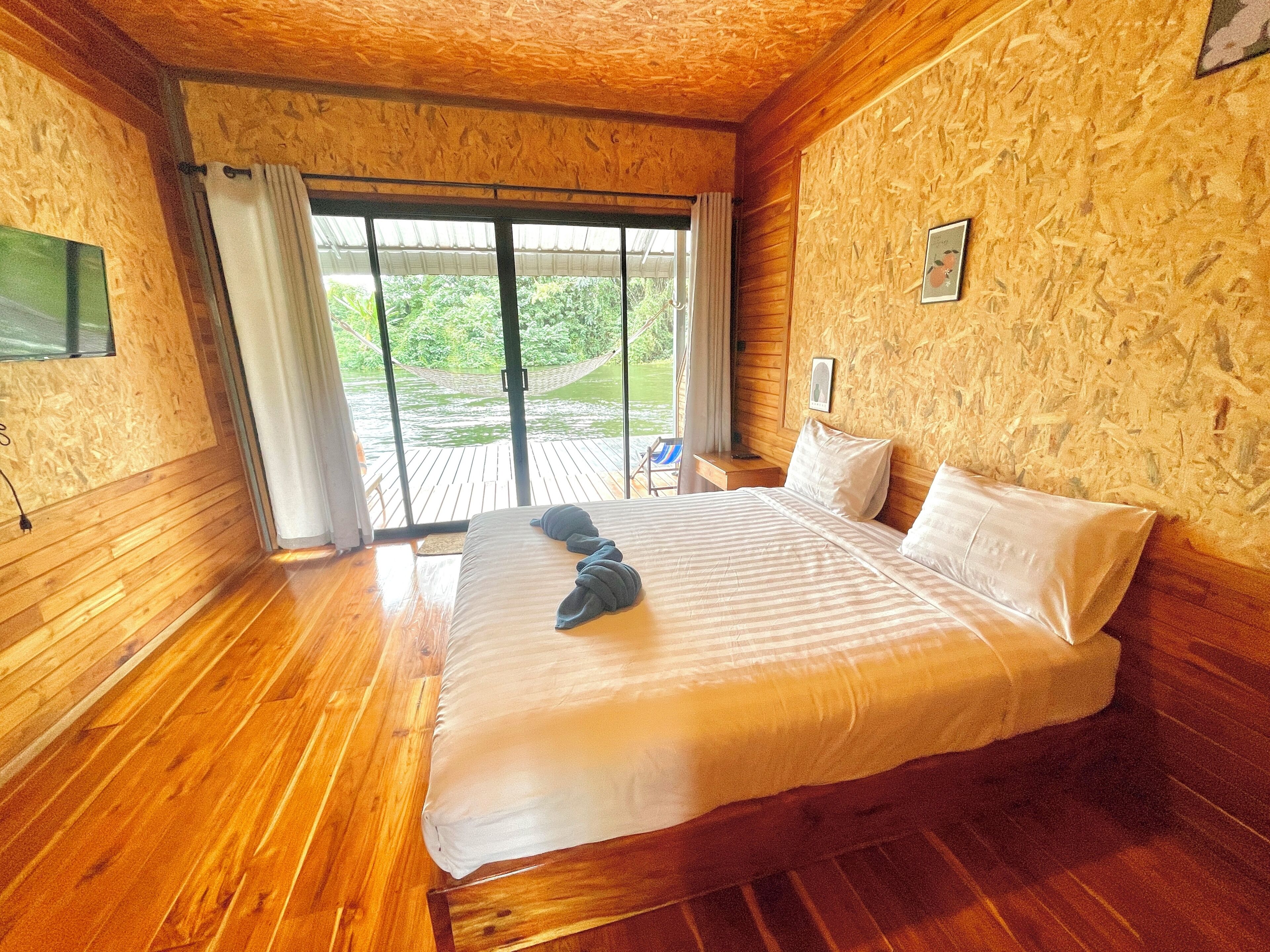 A bedroom with a large bed, wooden flooring, a window with curtains, and a view of greenery outside.
