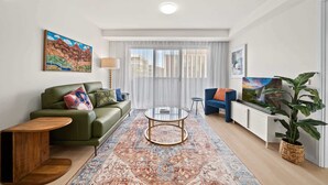 Un salon moderne avec un canapé vert, un fauteuil bleu, une table basse en verre et un grand tapis. Deux tableaux ornent les murs, ainsi qu’une lampe et une plante en pot.