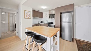 Une cuisine moderne avec des meubles en bois, un réfrigérateur en acier inoxydable, un four à micro-ondes et une table à manger en bois, accompagnée de deux tabourets noirs.