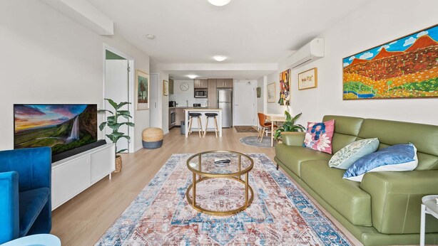 Un salon moderne avec un canapé vert, une table basse et un téléviseur. On aperçoit, au fond, une cuisine équipée d’un réfrigérateur et d’un four à micro-ondes.